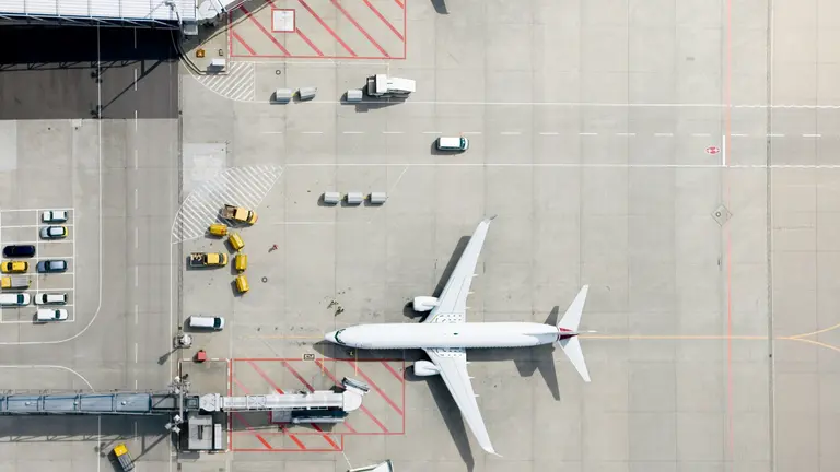 Aerial view of a commercial aircraft parked at an airport gate, surrounded by ground service vehicles, equipment, and boarding bridges on the apron.