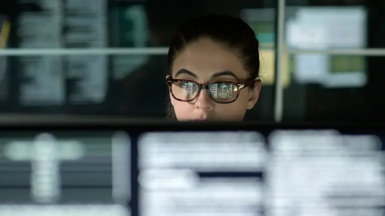 A woman with glasses sitting behind  computer monitors