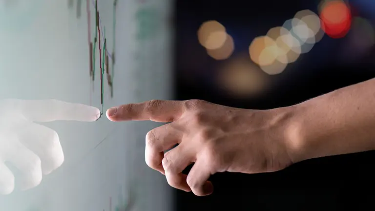 A hand pointing on a monitor, where stock prices can be seen