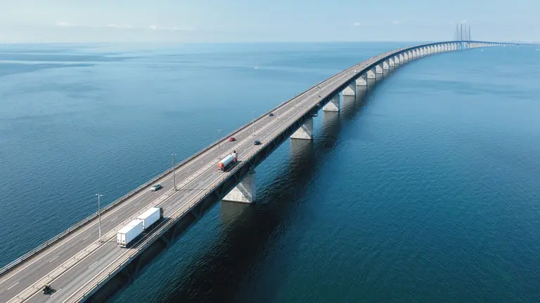 Aerial view of a long multi-lane bridge stretching across calm blue water, with several cars and trucks traveling along the roadway under a clear sky.