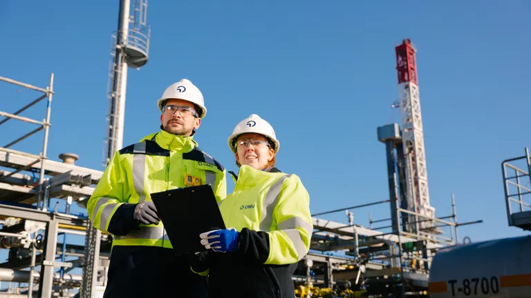 Two workers wearing safety equipment inspect operations at an industrial gas processing facility with pipelines and processing infrastructure in the background.