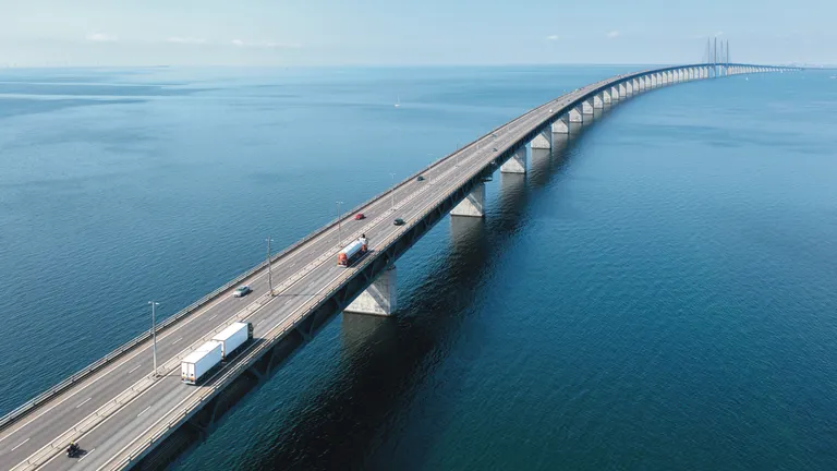 Aerial view of a long multi-lane bridge stretching across calm blue water, with several cars and trucks traveling along the roadway under a clear sky.