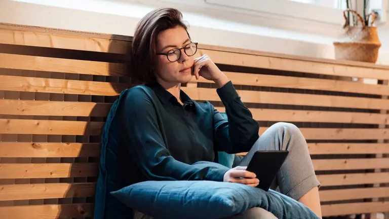 Woman sitting on a cushion against a wooden wall, reading on a tablet, deep in thought.