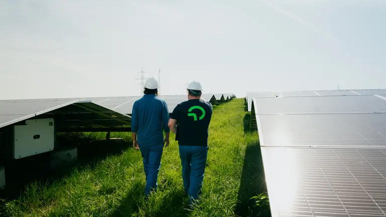 Two workers in safety helmets walk between rows of solar panels in a large solar energy installation on a sunny day.